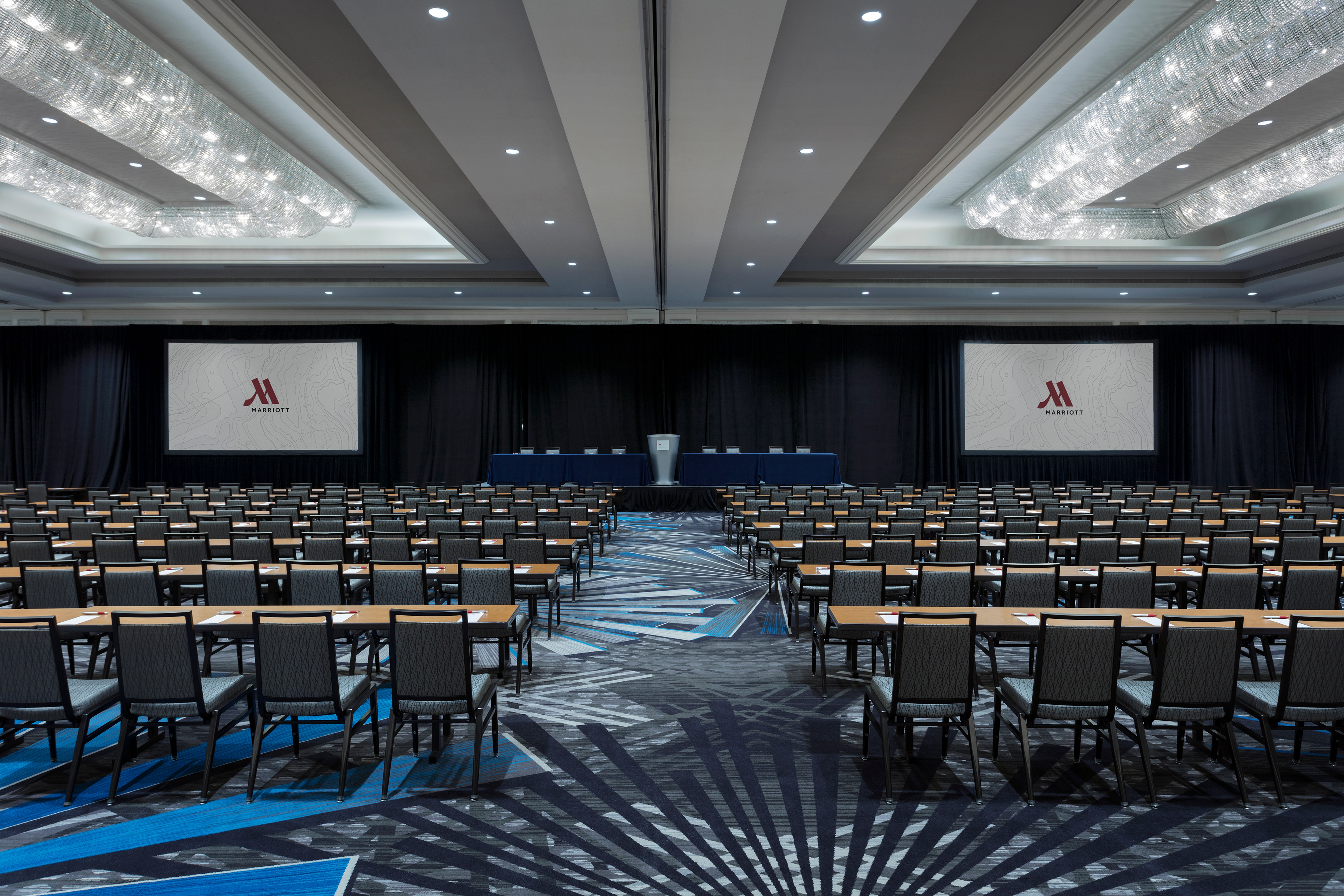 Carpeted ballroom with long rows of tables and chairs facing stage with podium