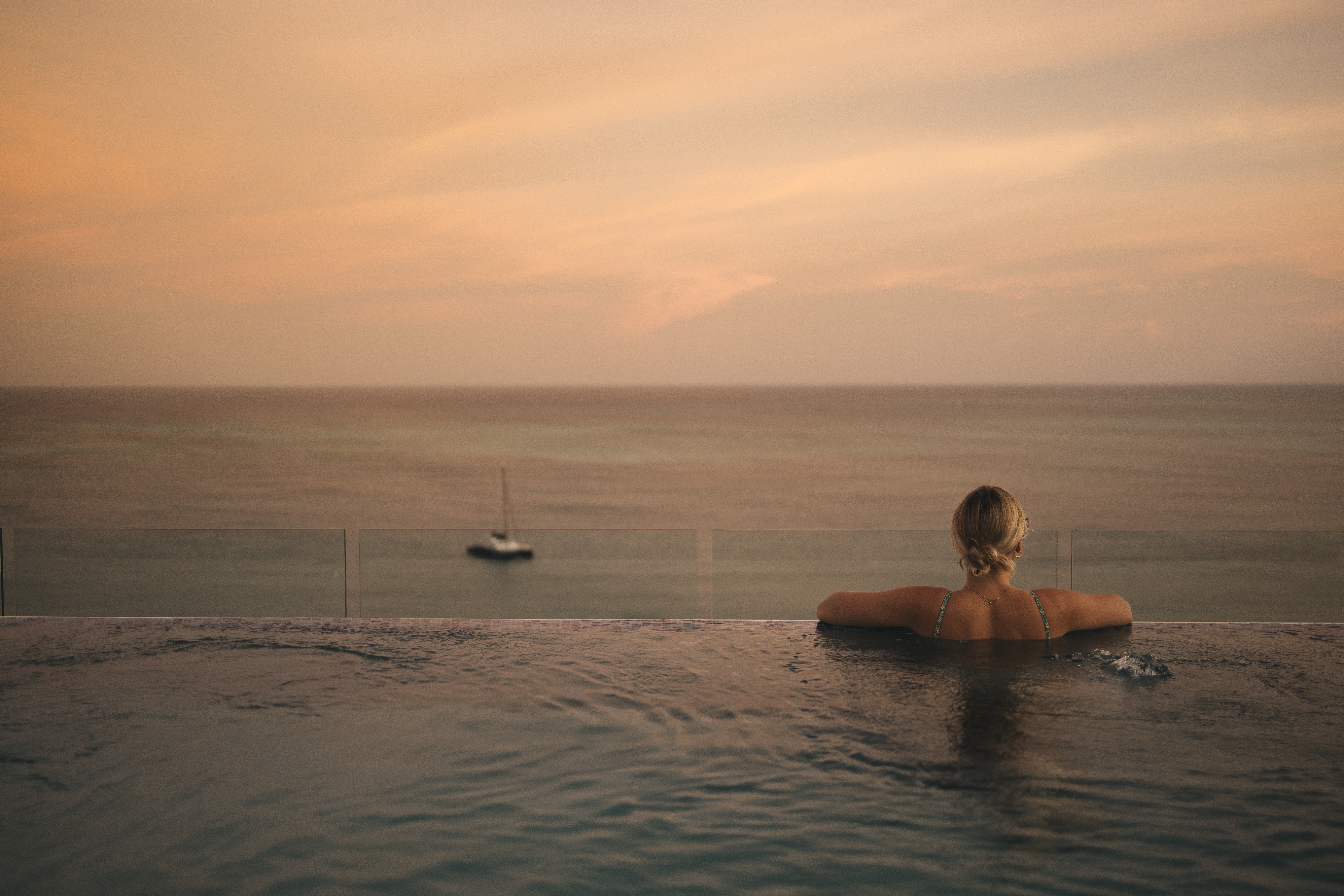 Mulher de costas na piscina olhando para o mar ao pôr-do-sol