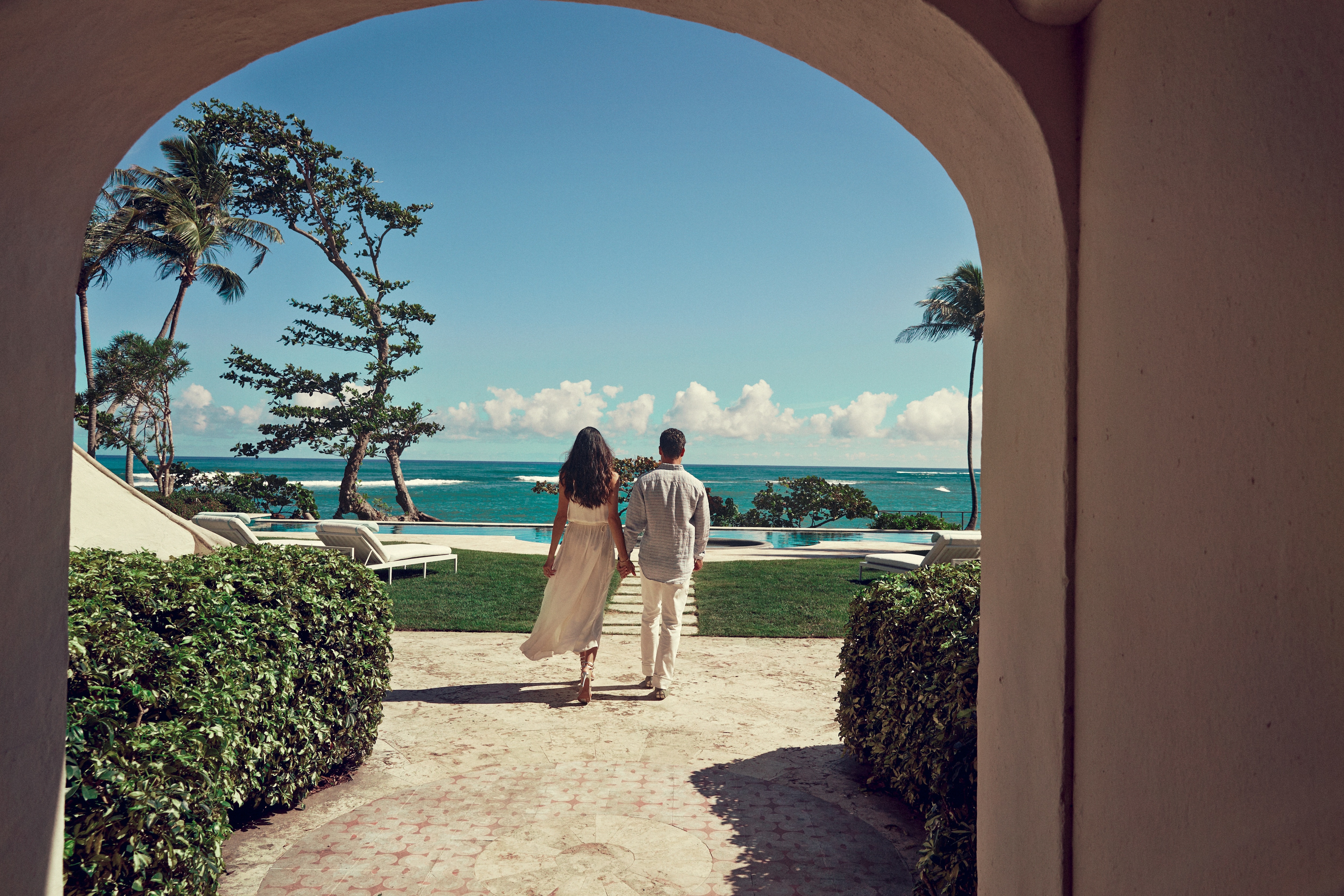 Um casal andando para trás no exterior do hotel com o mar ao fundo.