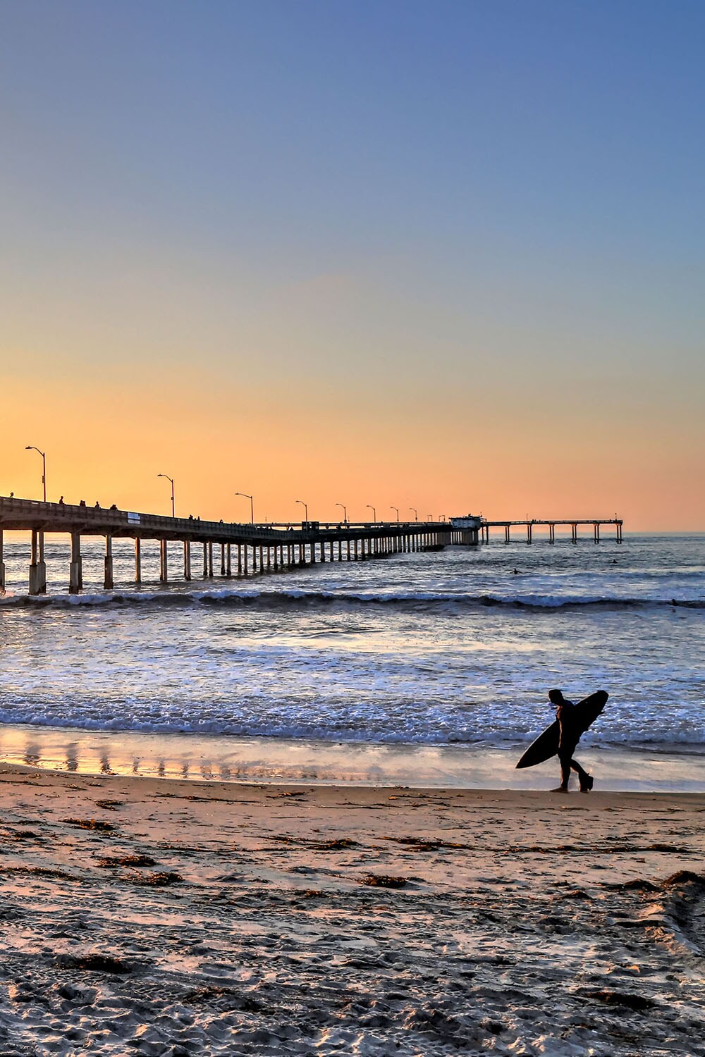 surfer walking on a beach with a pier in the background
