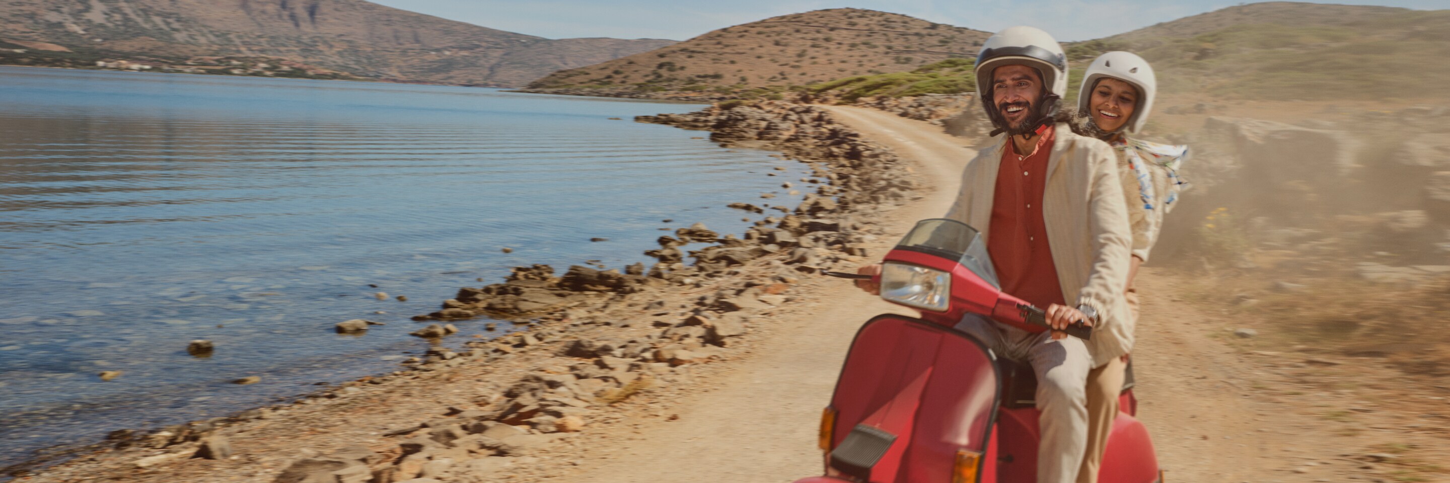 A couple rides a scooter on a dirt road near a body of water