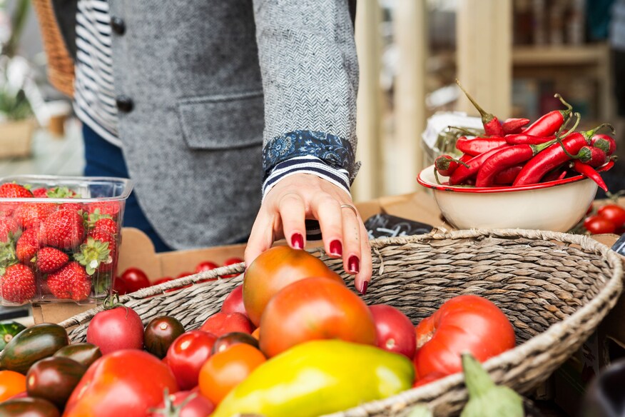 A well-manicured hand with red fingernails reaches into a woven bowl of red tomatoes at a market