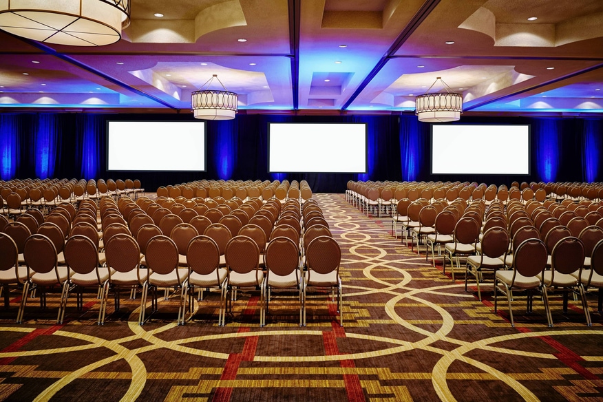 Large ballroom with hundreds of seats facing a lecturn and three large screens.
