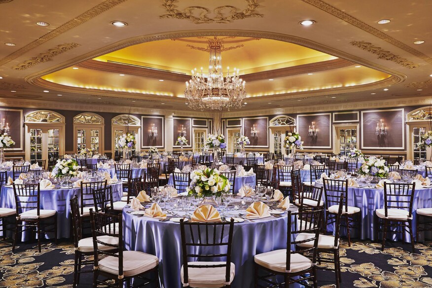 Luxurious ballroom with chandelier, paintings, and round tables with flower arrangements and elaborate table settings.
