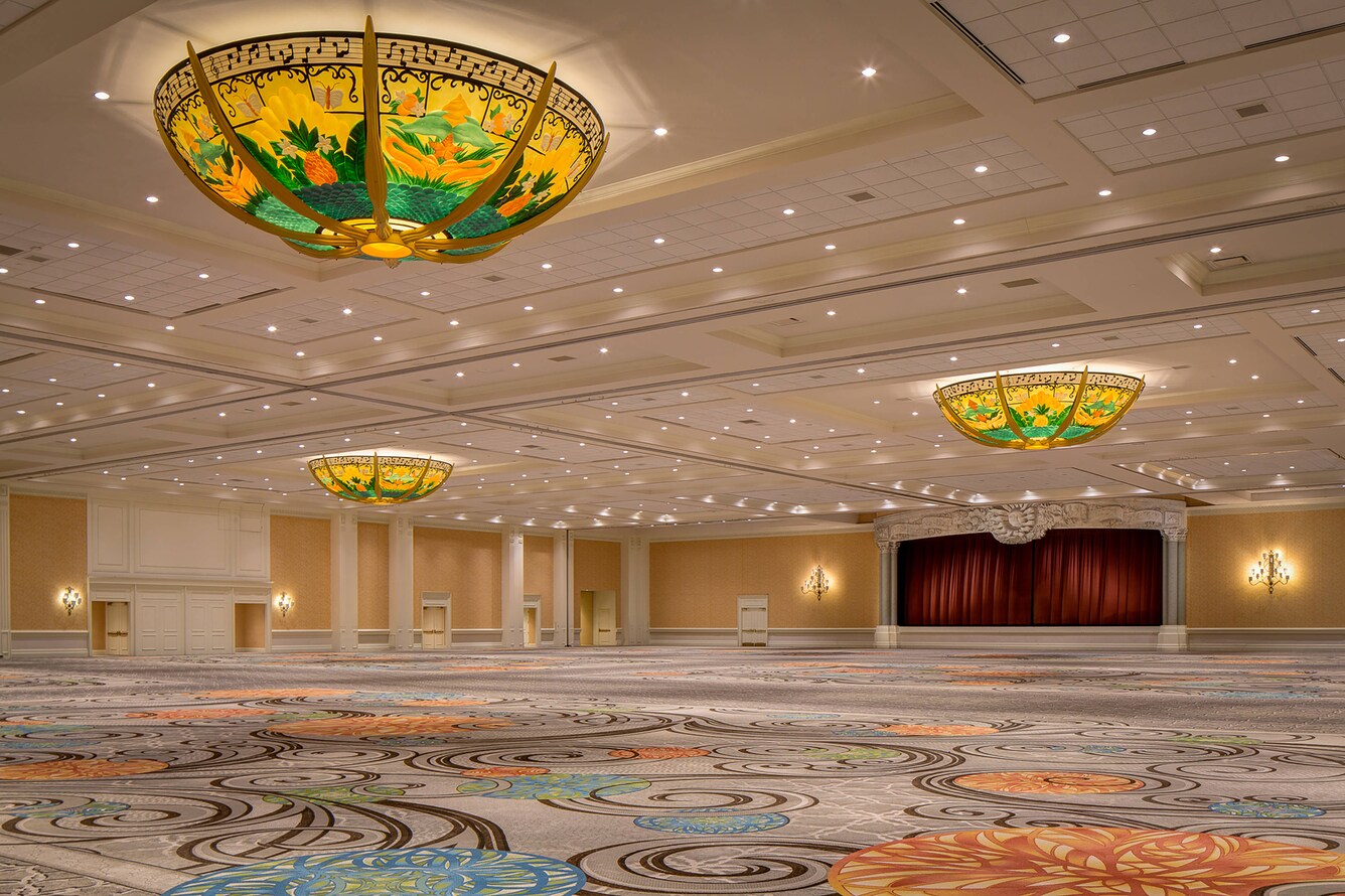 View of empty ballroom space with patterned carpet, stage, and colorful chandelier.