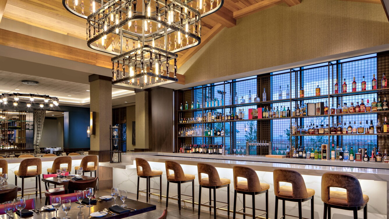 Restaurant dining area seating, bar, barstools and bottles of liquor illuminated by a chandelier.