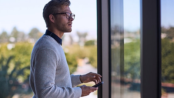 Man with a cell phone gazing out a tinted window of an office building.