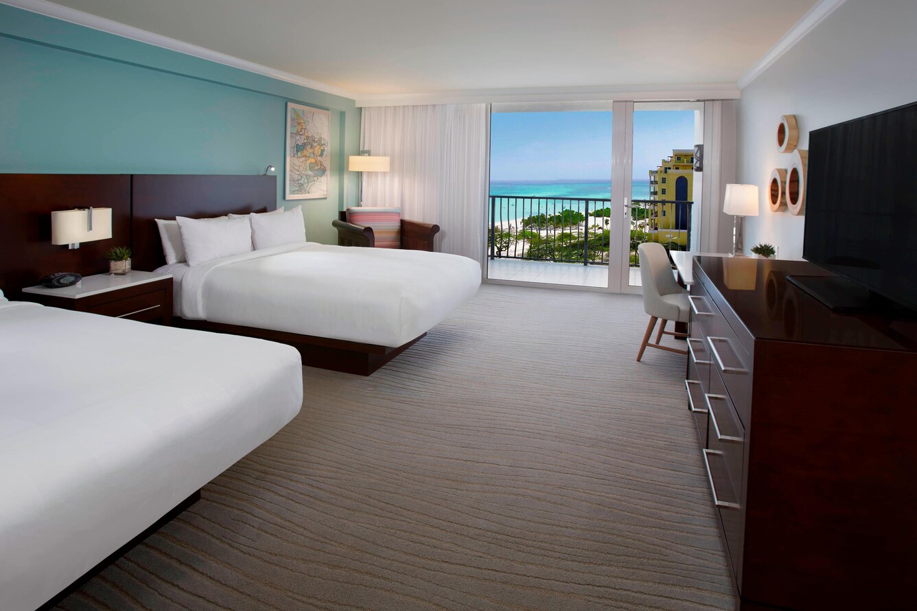 Guestroom with two queen beds and a partial view of the balcony, beach, ocean, and trees.