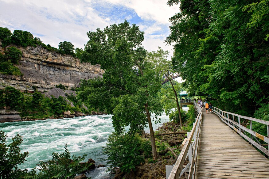 Wooden slat walkway alongside a whitewater river canyon with lush, green trees.