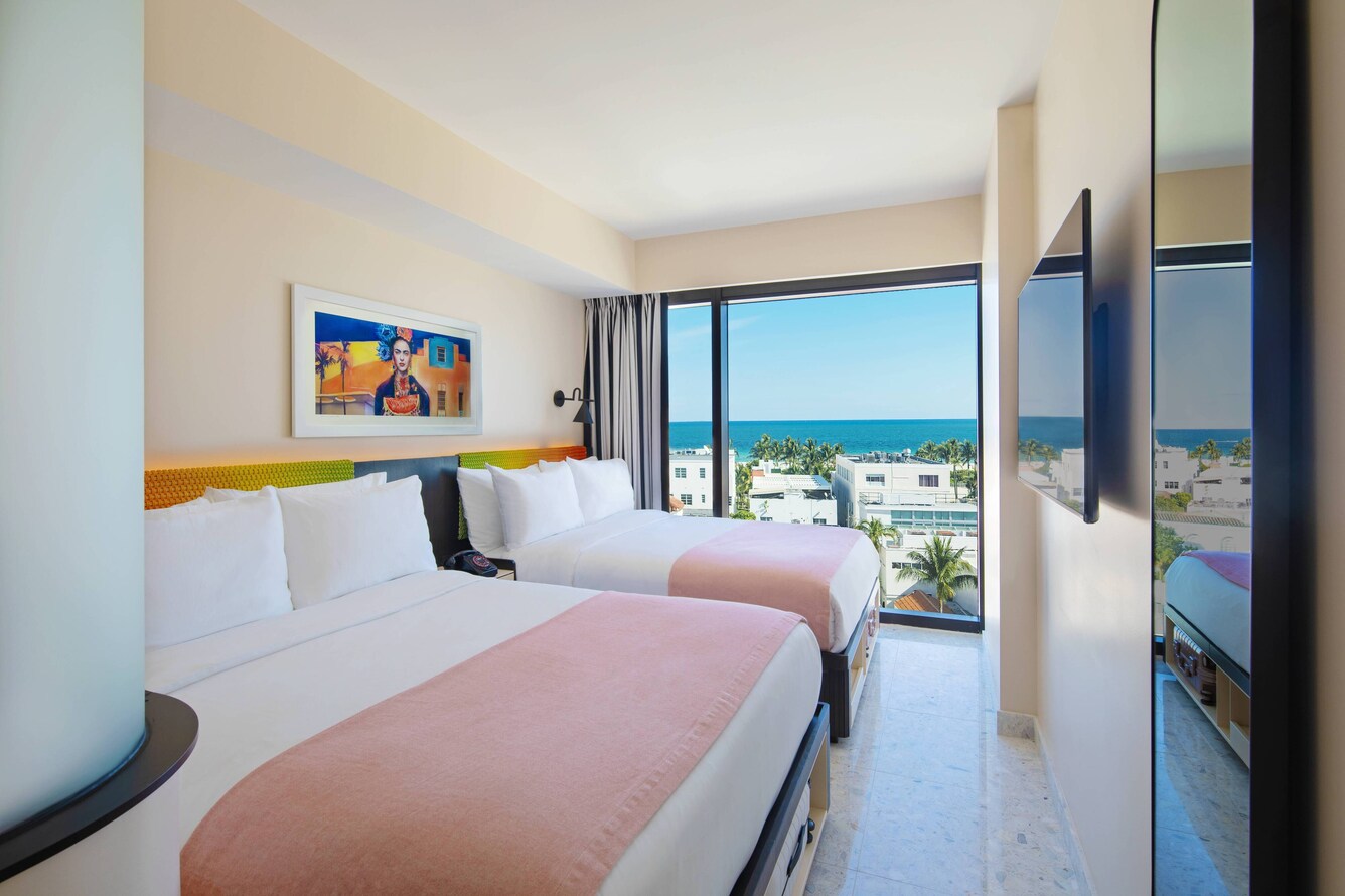 Two queen beds with pink coverlets, multicolored art, and views of the ocean