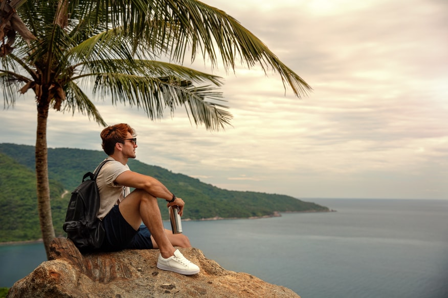 Man sitting on island under palm tree staring at the sea at sunset.