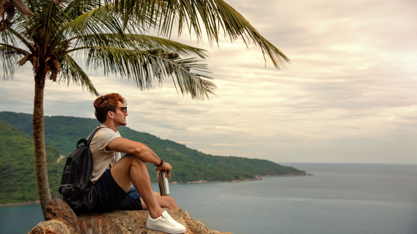 Man sitting on a hill under a palm tree overlooking the ocean.
