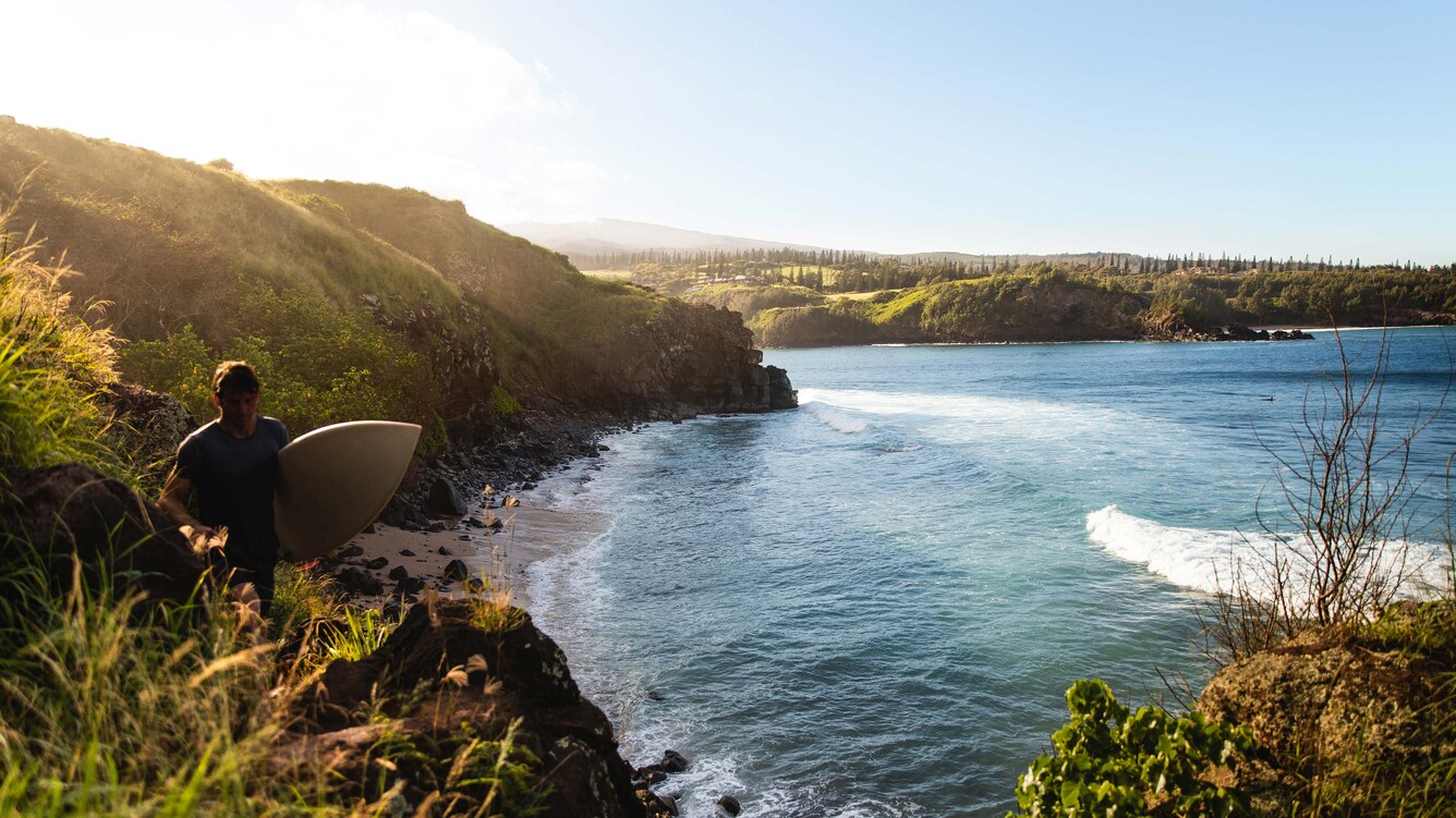 Man with a surfboard under his arm walking along the coastal cliffs.