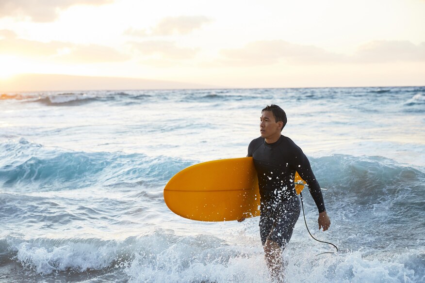 Man in a wetsuit with a surfboard under his arm walking in from the surf.