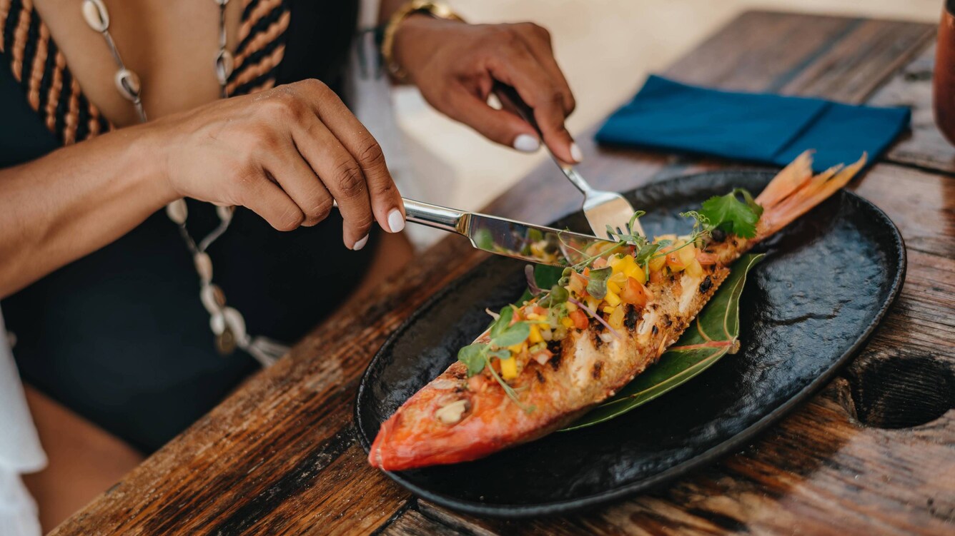 Woman's hands cutting up a whole red snapper with a fork and butter knife.