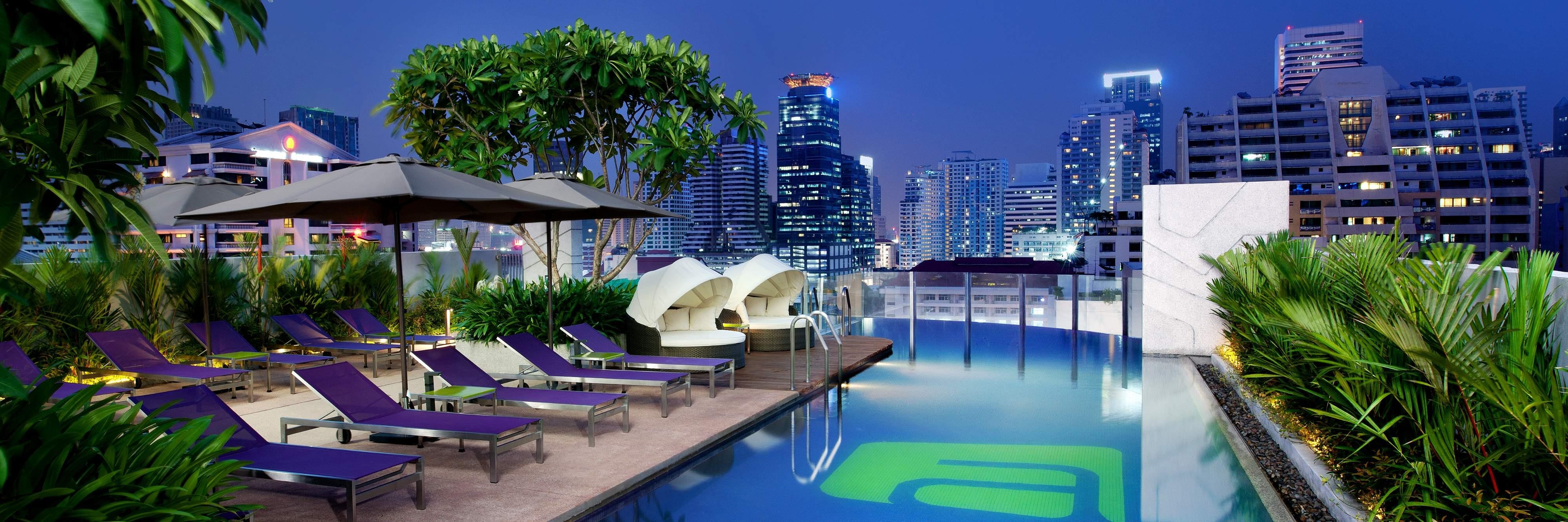 Rooftop swimming pool surrounded by deck chairs and plants with city view