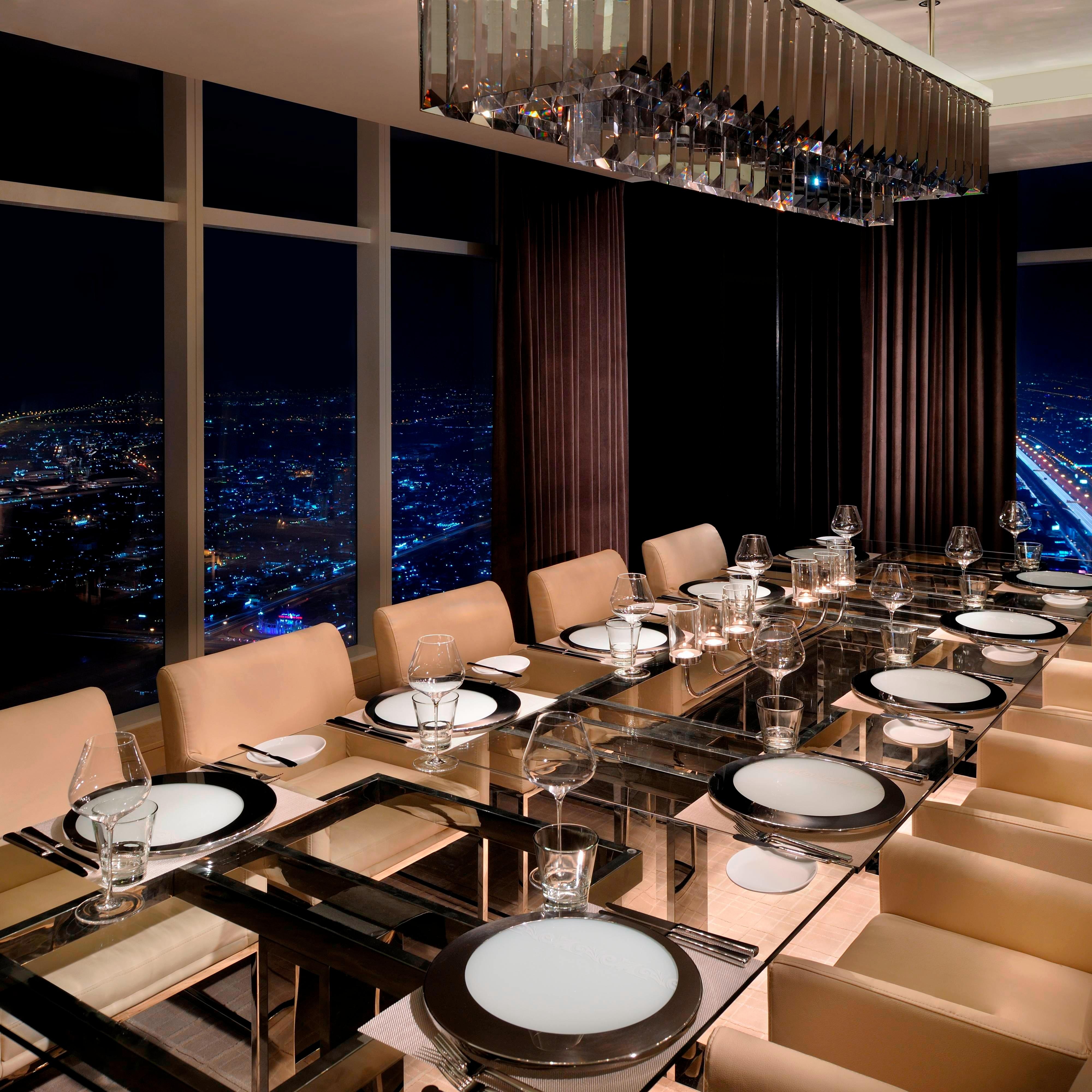 A large dining table set beside windows with city views at night