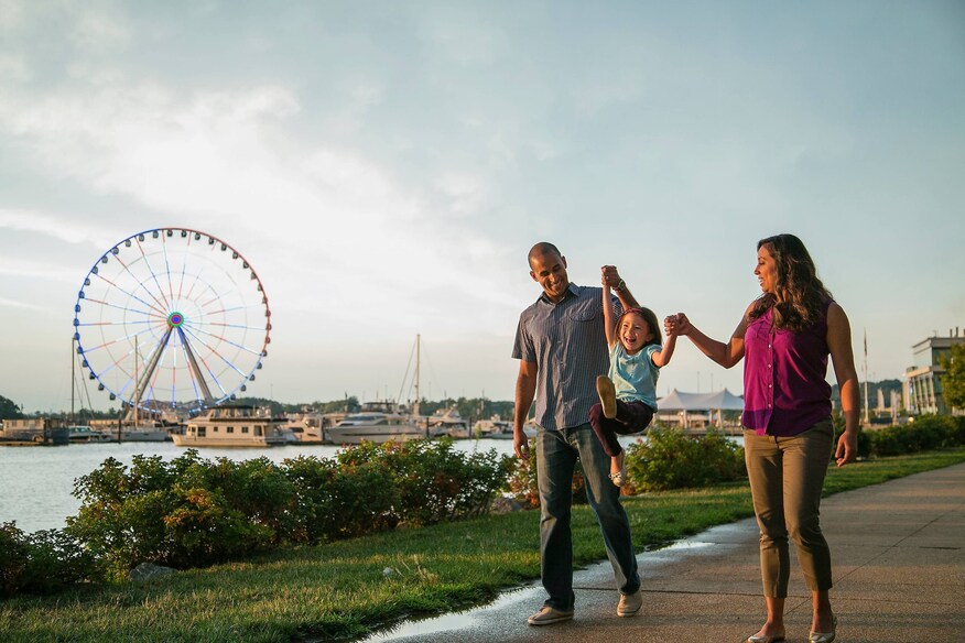Man, woman, and child holding hands and walking on harbor path with a ferris wheel in the background.