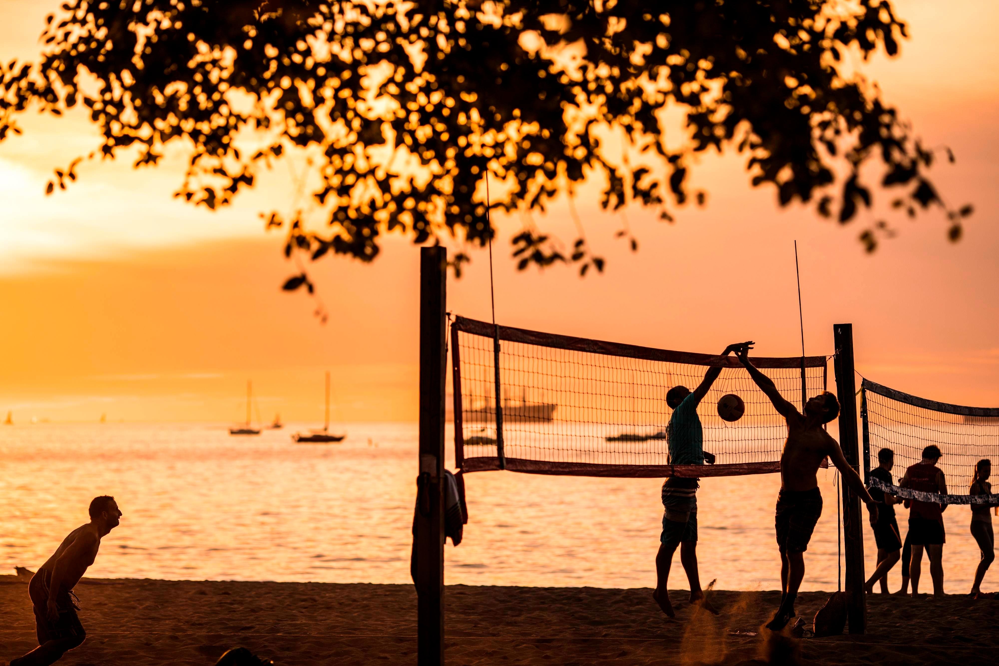 People playing volleyball outdoors, sillhouetted by the sun setting over the water.