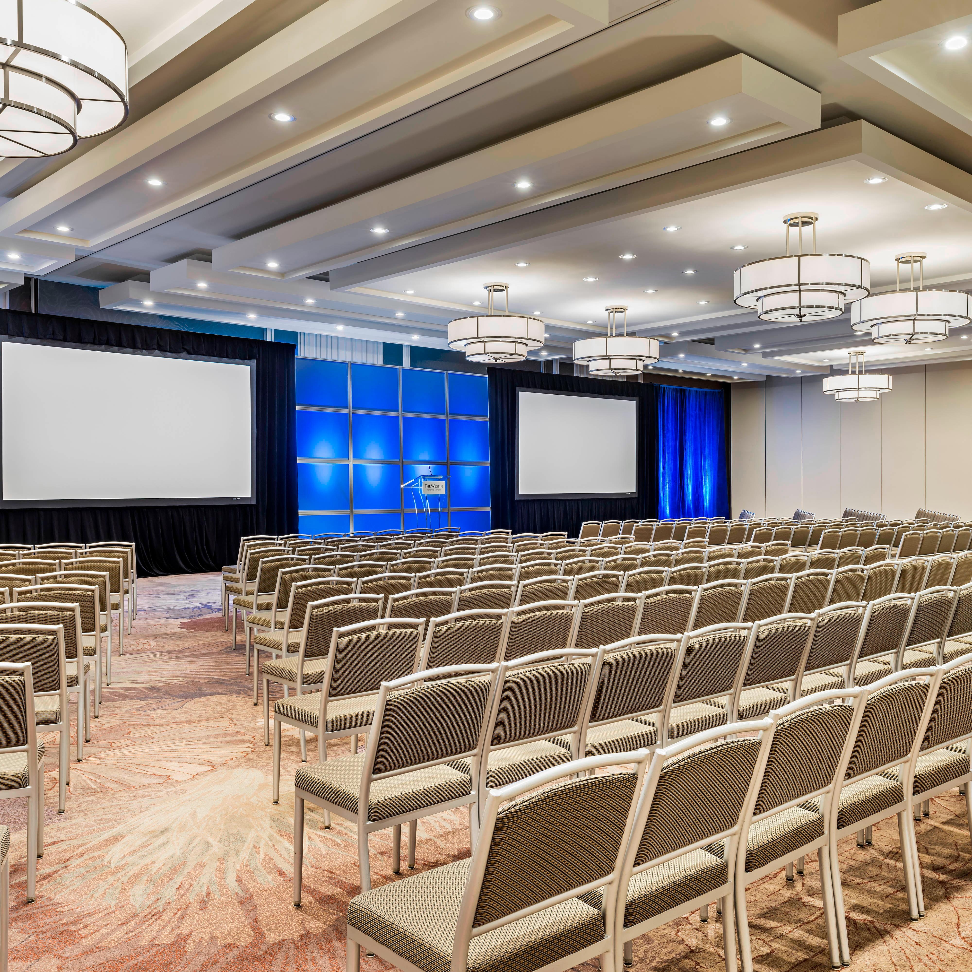 Ballroom event setup with rows of chairs and blue-lit stage with projector screens